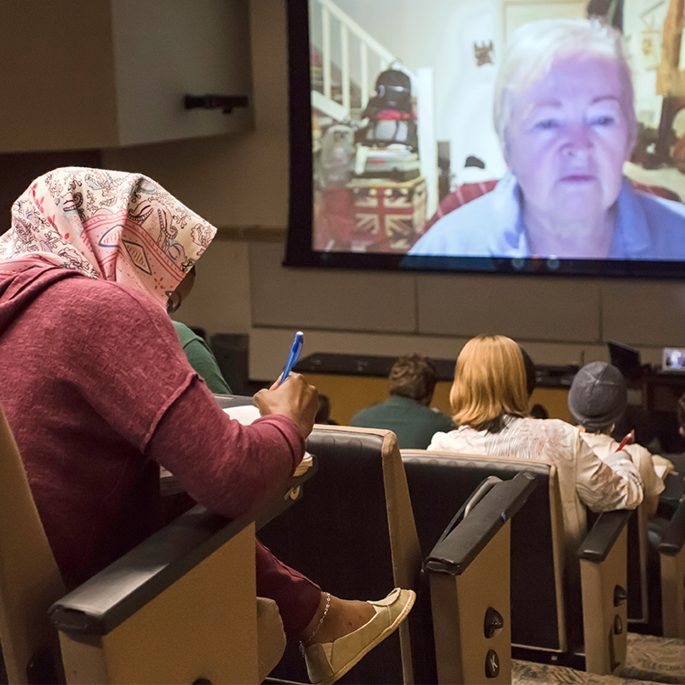 Students take notes in an auditorium while Skype video of anime expert Helen McCarthy is projected on a screen at the front of the classroom.